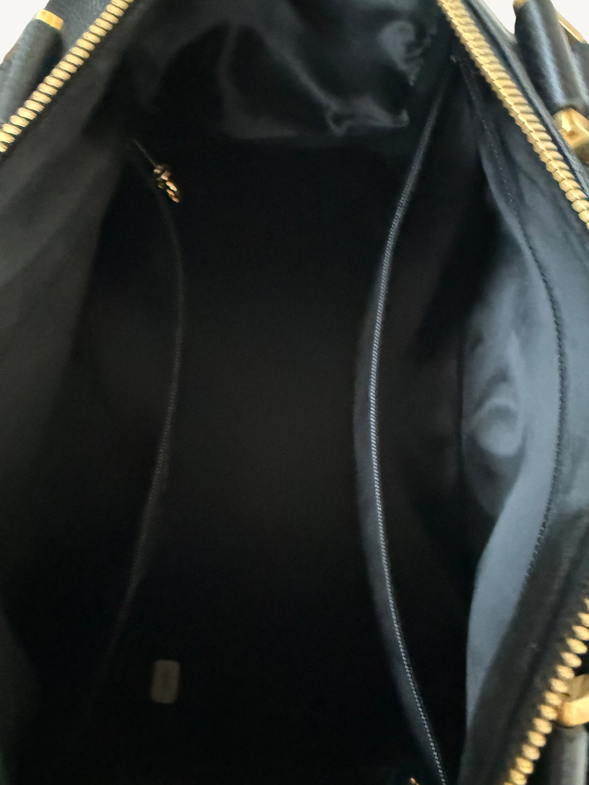 Close-up view of the interior of a black handbag, showing a black fabric lining, two central zip compartments, and gold-tone zipper hardware.