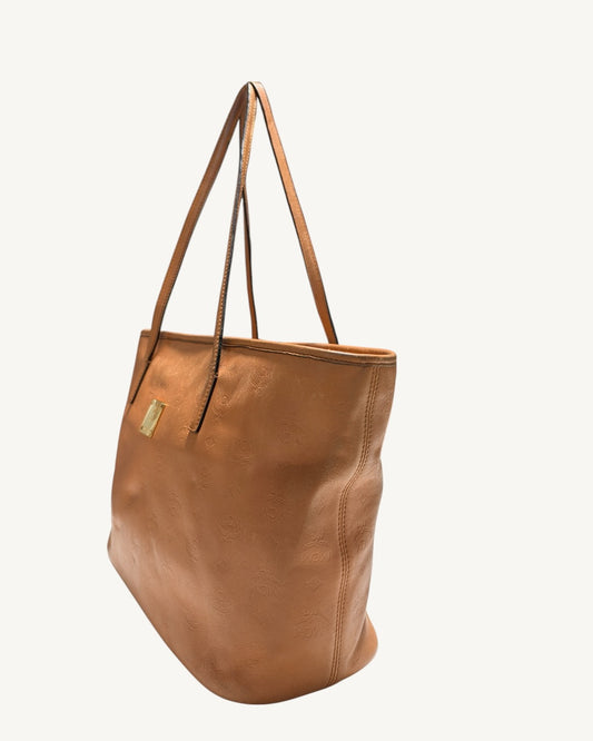MCM cognac tote angled view showing the right side seam, front plaque and long leather straps.