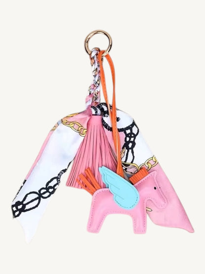 A pink leather unicorn keychain charm featuring a light blue wing, orange mane, and a pink tassel, attached to a patterned white scarf with black chains and gold lettering, all on a gold key ring.