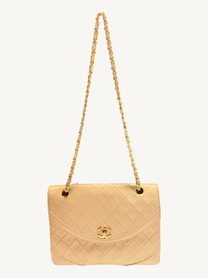 A  beige quilted Chanel leather shoulder bag with a gold-tone chain strap and a gold interlocking CC logo on the front flap, set against a plain off-white background.