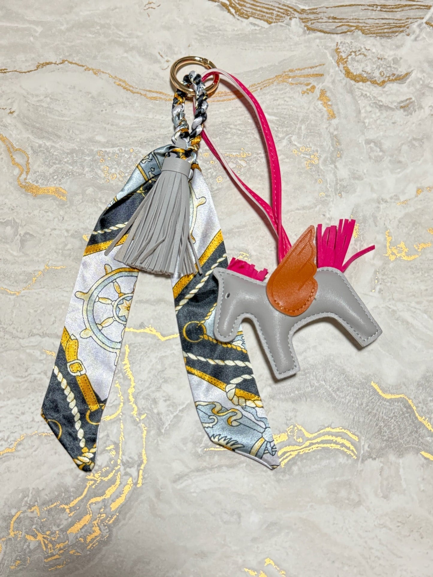 A decorative keychain or bag charm laid flat on a light marble-like surface. The set includes a gray horse-shaped leather figure with a tan saddle, a bright pink strap and tail, a gray tassel, and a patterned scarf with nautical-style motifs in gray, white, yellow, and blue attached to a metal ring.