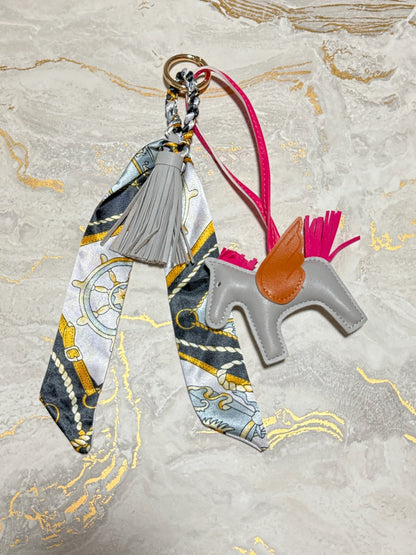 A decorative keychain or bag charm laid flat on a light marble-like surface. The set includes a gray horse-shaped leather figure with a tan saddle, a bright pink strap and tail, a gray tassel, and a patterned scarf with nautical-style motifs in gray, white, yellow, and blue attached to a metal ring.