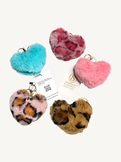 Five fuzzy faux-fur heart-shaped keychains in different colors and patterns, including turquoise, dusty rose, light pink, leopard print, and tan leopard print, arranged on a light cream background with two branded tags visible.