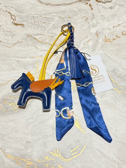A designer keychain charm featuring a blue leather dalmatian dog silhouette with a brown leather saddle, attached to a blue silk scarf with gold chain print, a blue tassel, and a yellow strap, displayed on a white marble background with gold veins. A white tag for 'Summer Shawls' is visible.