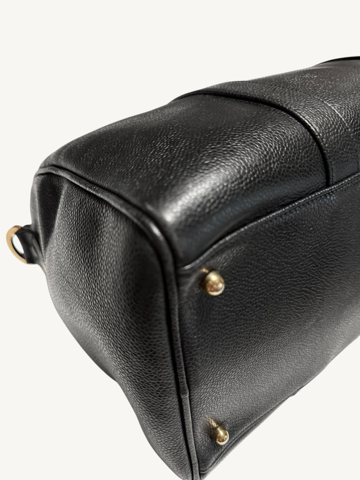 Close-up of a black textured leather handbag corner with small gold-tone metal studs and a partial gold ring detail, photographed on a clean white background.