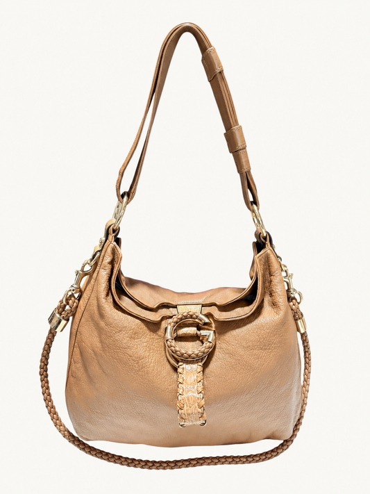 A camel brown Gucci leather shoulder bag with a large circular G gold-tone front harware, a woven strap detail, and a braided shoulder strap, shown against a plain off-white background.