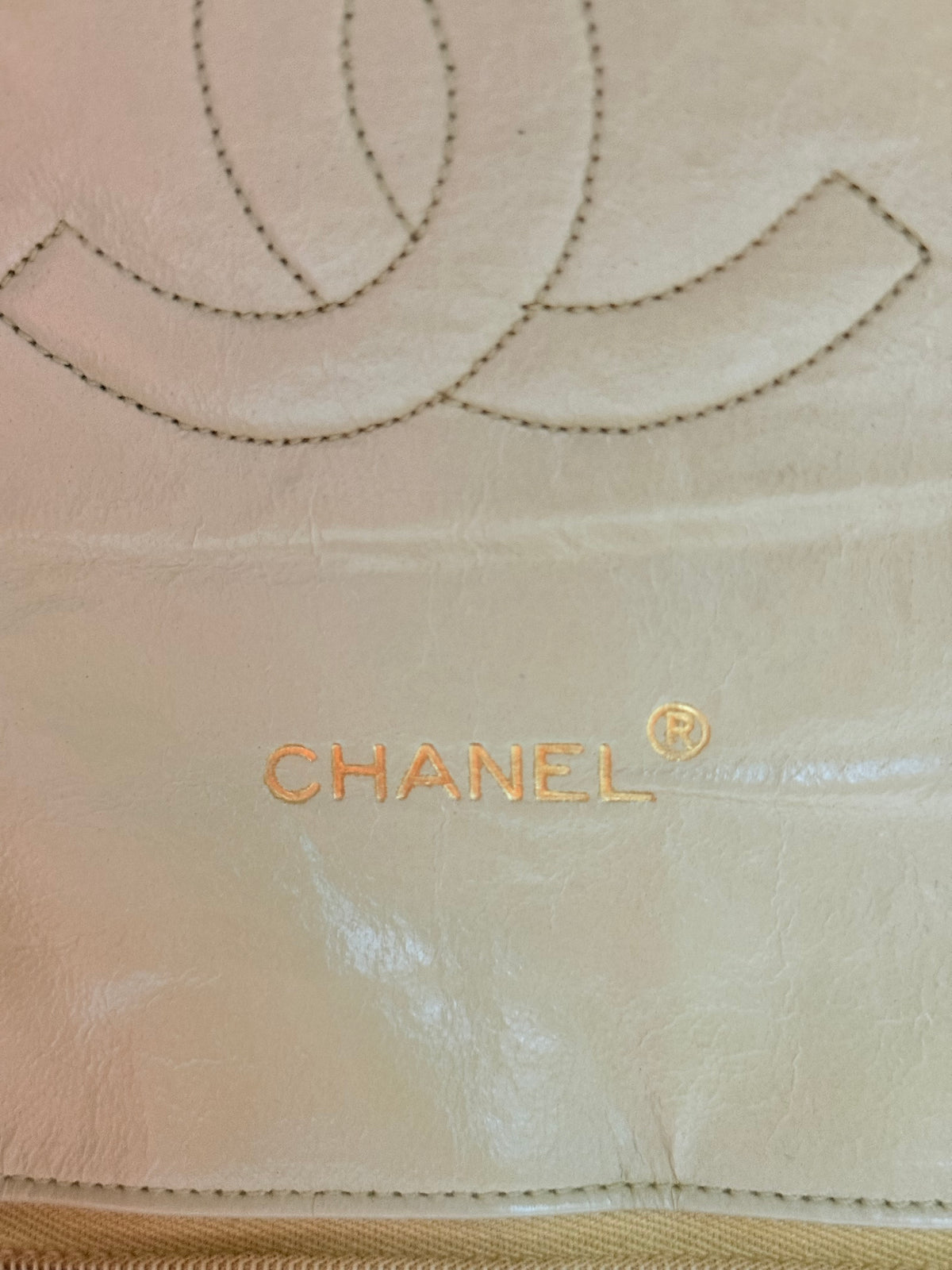 Close-up of a Chanel bag in light beige leather, showing the interlocking CC logo and CHANEL text in gold-tone embroidery.