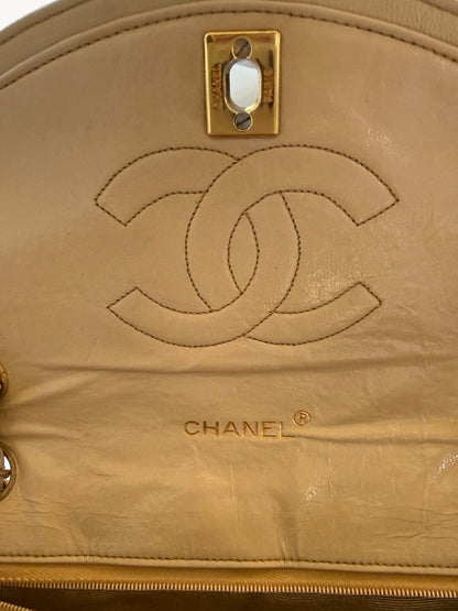 Close-up of a beige leather Chanel handbag showing the embossed interlocking CC logo, CHANEL text, a gold-tone metal clasp, and part of the zipper.