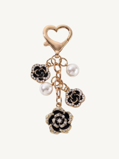 A gold-tone heart-shaped clasp keychain or bag charm featuring three dangling rose charms. Two of the rose charms are black with white pearl accents, and one larger rose charm at the bottom is black with clear rhinestone detailing.