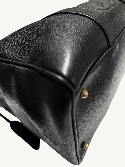 Close-up of a black pebbled leather handbag corner showing a gold-tone metal stud and a black leather strap against a light off-white background.