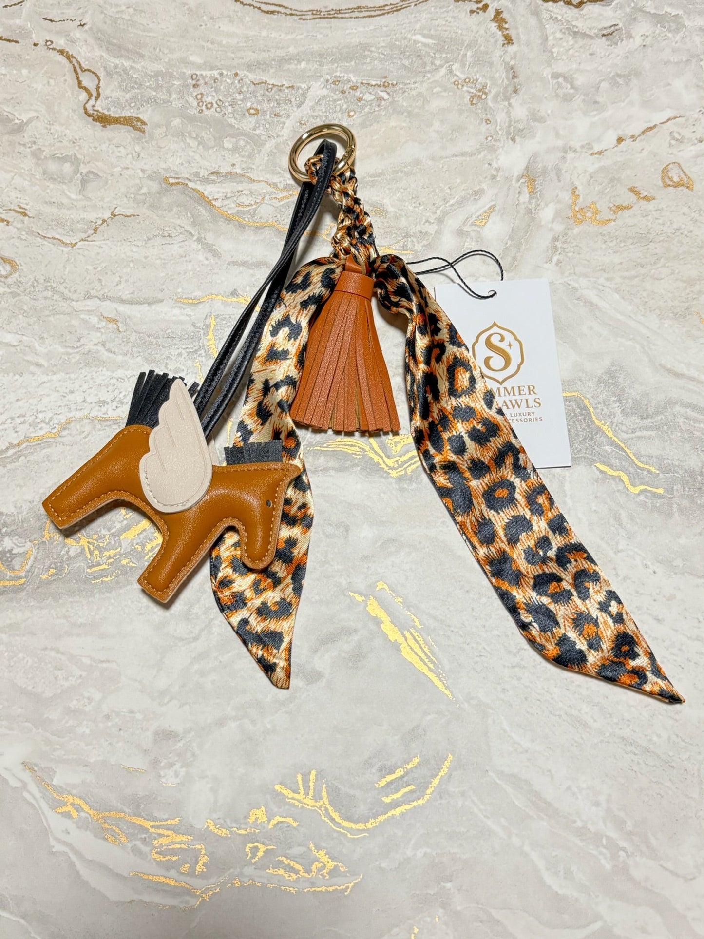 A horse-shaped leather keychain charm with a leopard-print ribbon, a brown leather tassel, and a white brand tag reading 'Saver Axles' on a light marble background.
