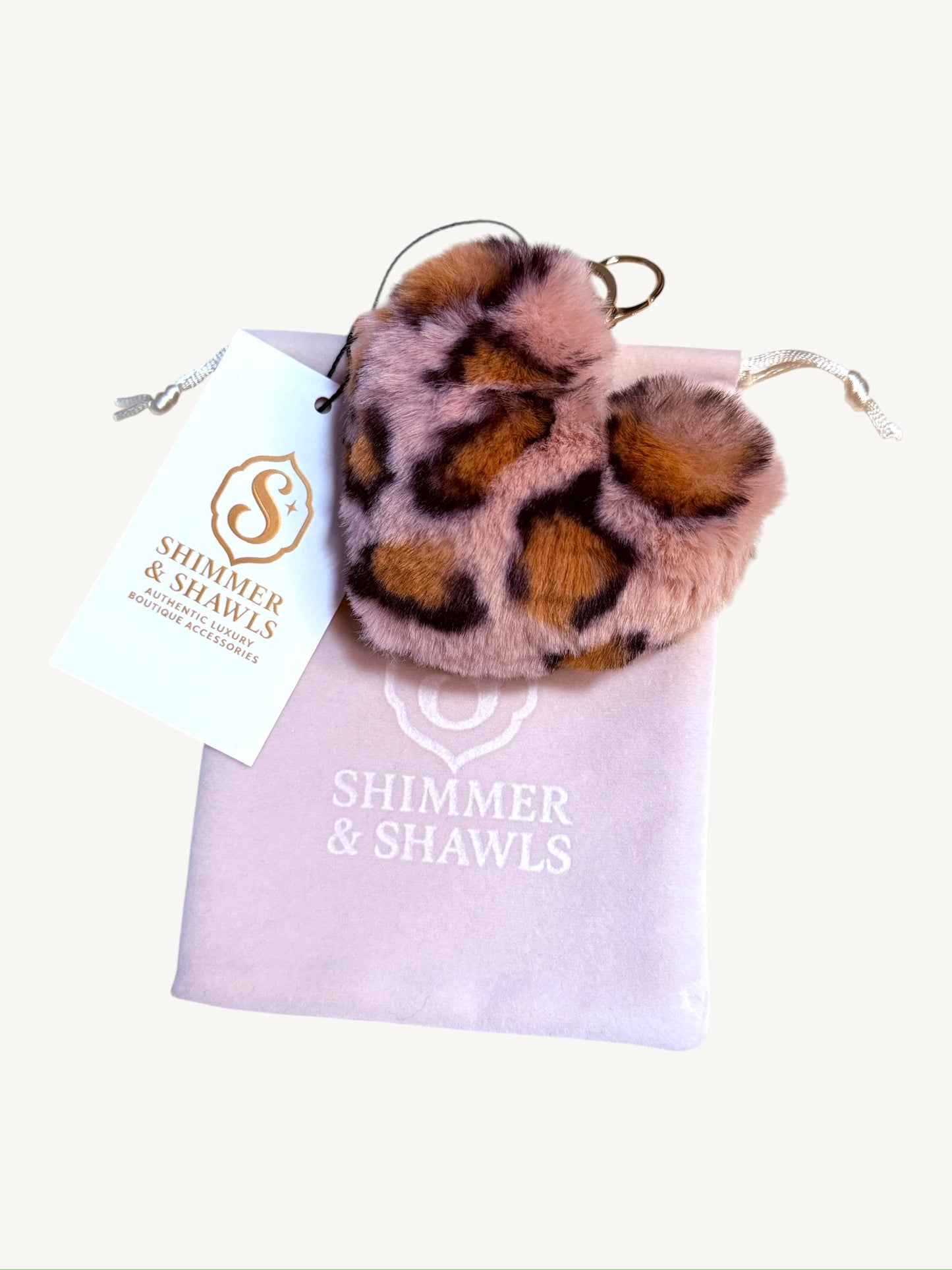A leopard-print faux fur pom pom keychain or bag charm with a gold metal ring, presented on top of a light pink drawstring bag with a white 'Shimmer & Shawls' brand tag.