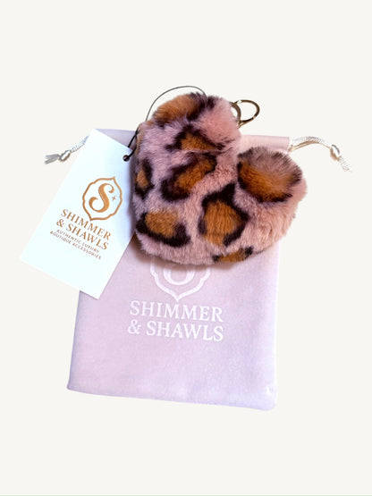 A leopard-print faux fur pom pom keychain or bag charm with a gold metal ring, presented on top of a light pink drawstring bag with a white 'Shimmer & Shawls' brand tag.