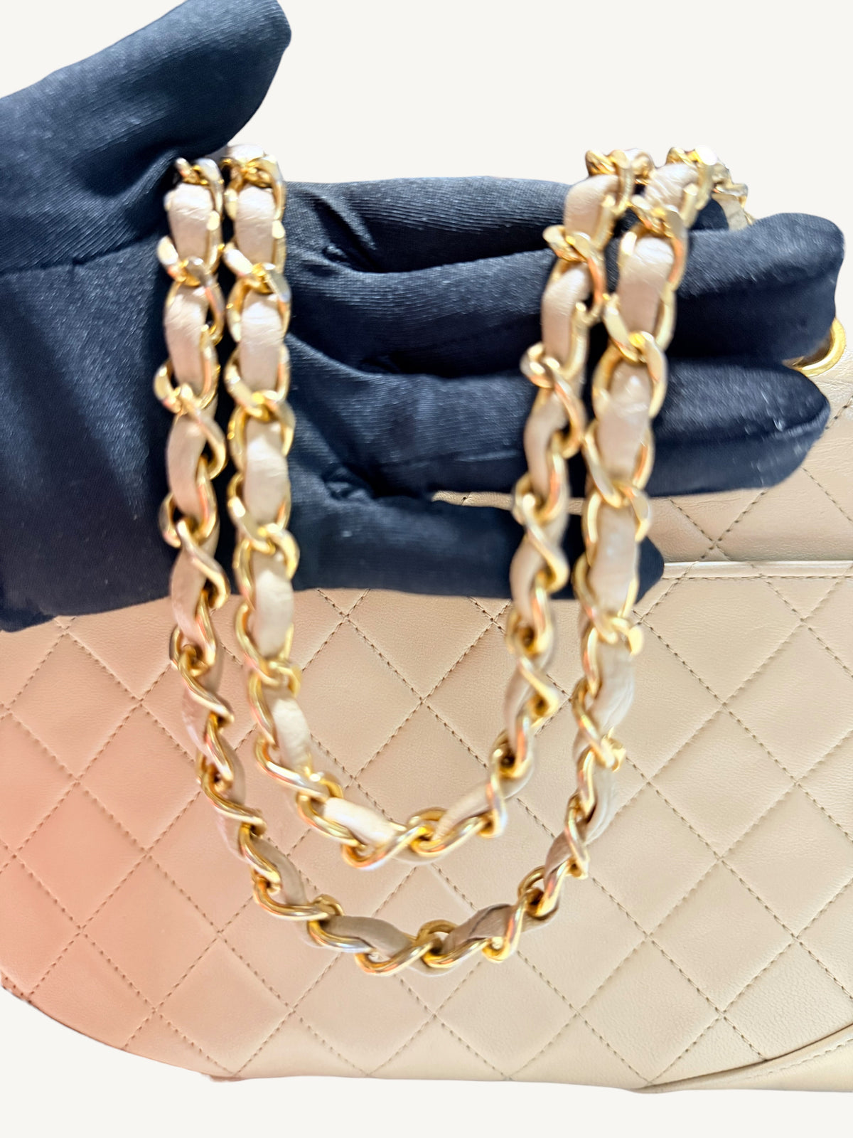 A close-up of a quilted handbag in light beige leather with a double chain strap featuring gold-tone hardware, held by a dark blue satin-gloved hand against a light background.