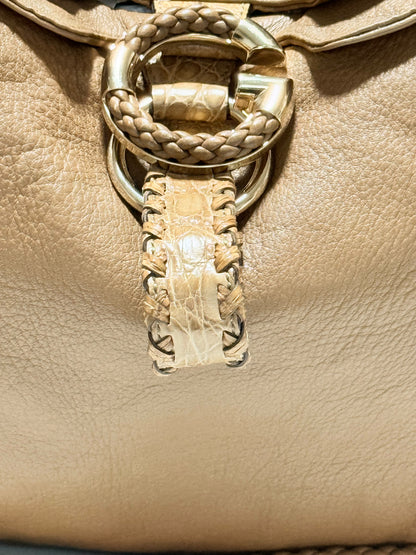 Close-up of a camel brown Gucci leather handbag featuring a large circular gold-tone hardware ring with braided and crocodile-embossed strap details.