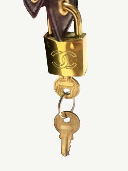 A close-up of a gold-toned padlock engraved with the interlocking CC logo, attached to a key ring with a key and a dark brown leather strap on a light gray background.