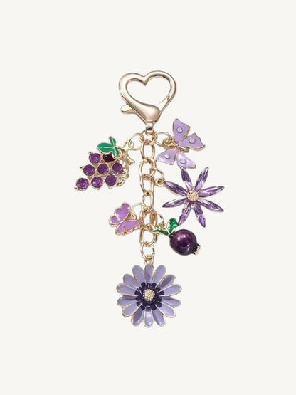A gold-tone charm keychain featuring purple enamel flowers, a grapevine cluster, and a purple berry, attached to a heart-shaped clasp on a light cream background.