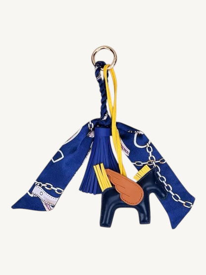 A decorative keychain charm featuring a navy blue silk scarf with white and gold chain motifs, a navy blue leather horse-shaped ornament, a brown wing charm, yellow tassels, and a gold metal key ring, shown on a plain off-white background.