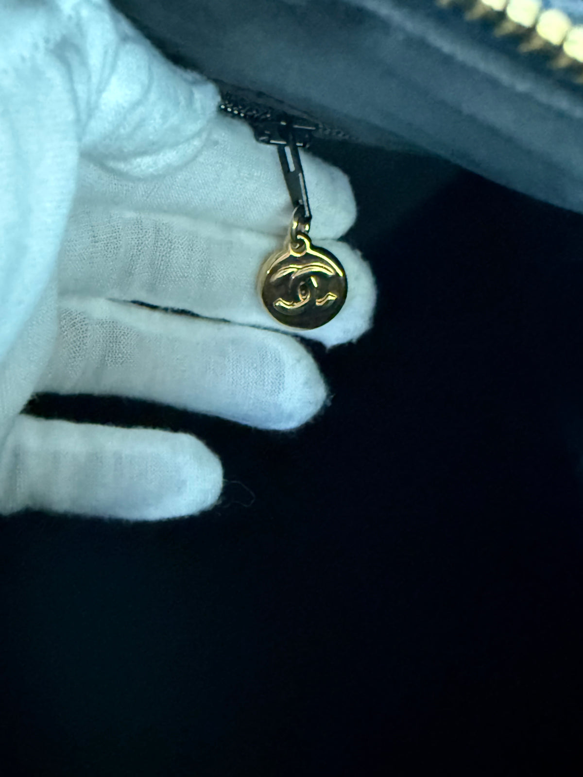 A close-up of a hand wearing a white glove holding a gold-toned circular zipper pull charm with a double-C logo, attached to a dark zipper.