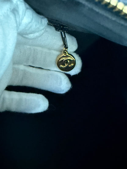 A close-up of a hand wearing a white glove holding a gold-toned circular zipper pull charm with a double-C logo, attached to a dark zipper.