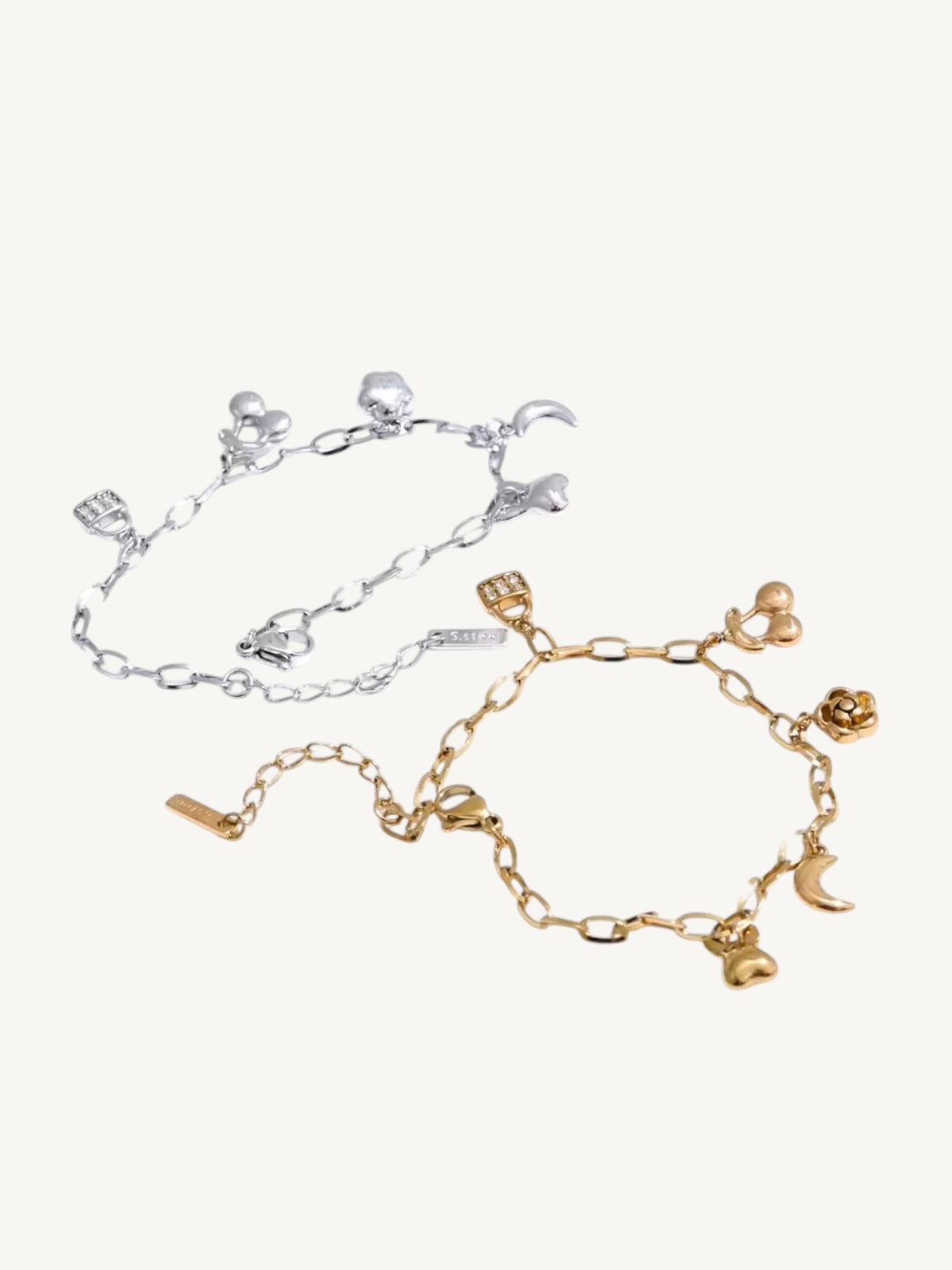 Two delicate chain bracelets with small charms, one in silver-tone metal and one in gold-tone metal, displayed on a light cream background.