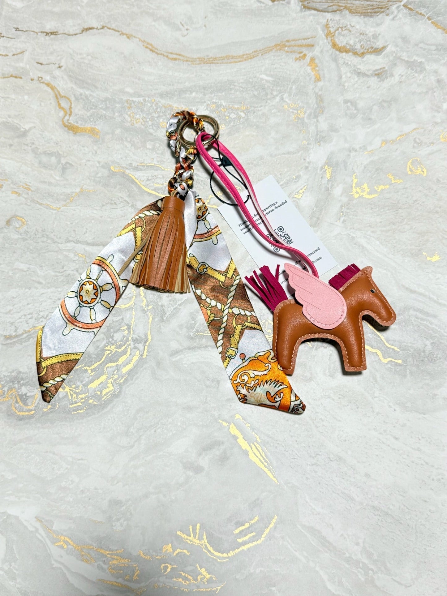 A luxury keychain bag charm featuring a brown leather horse with a pink saddle, a brown leather tassel, and a patterned silk scarf, displayed on a light marble background with gold veins.