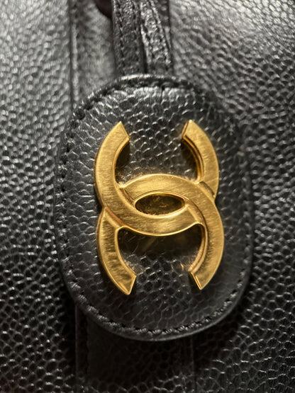 Close-up of a black textured leather bag featuring a prominent gold-tone interlocking double-C logo charm on an oval leather tab.