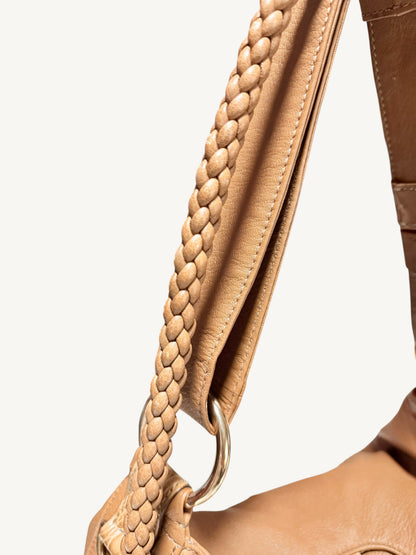 Close-up of a  camel brown Gucci leather shoulder bag showing a braided strap and a gold-tone metal ring on a light off-white background.
