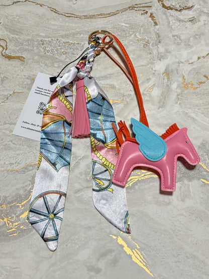A fashion keychain featuring a pink leather rocking horse charm with a light blue wing, attached to a patterned silk scarf, a pink tassel, and an orange wrist strap, displayed on a marble surface.
