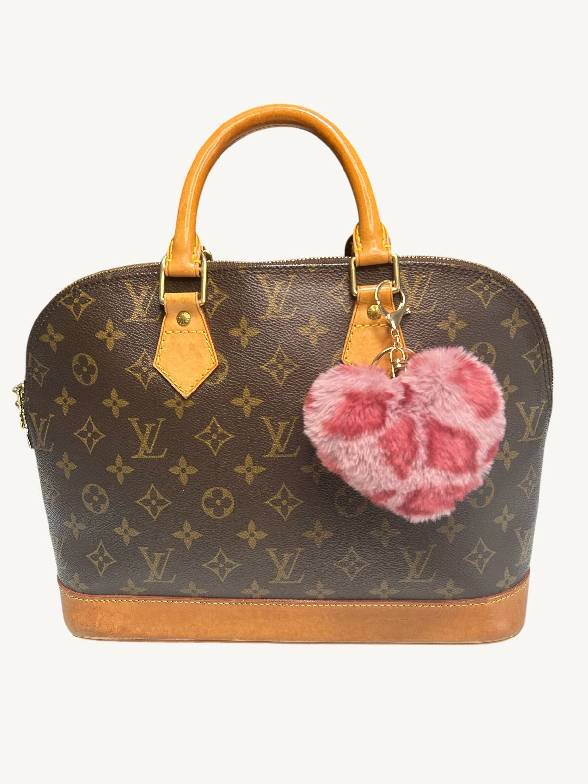 Louis Vuitton monogram handbag in brown coated canvas with tan leather handles and base trim, featuring a pink fuzzy heart-shaped charm hanging from the side, shown on a plain light background.