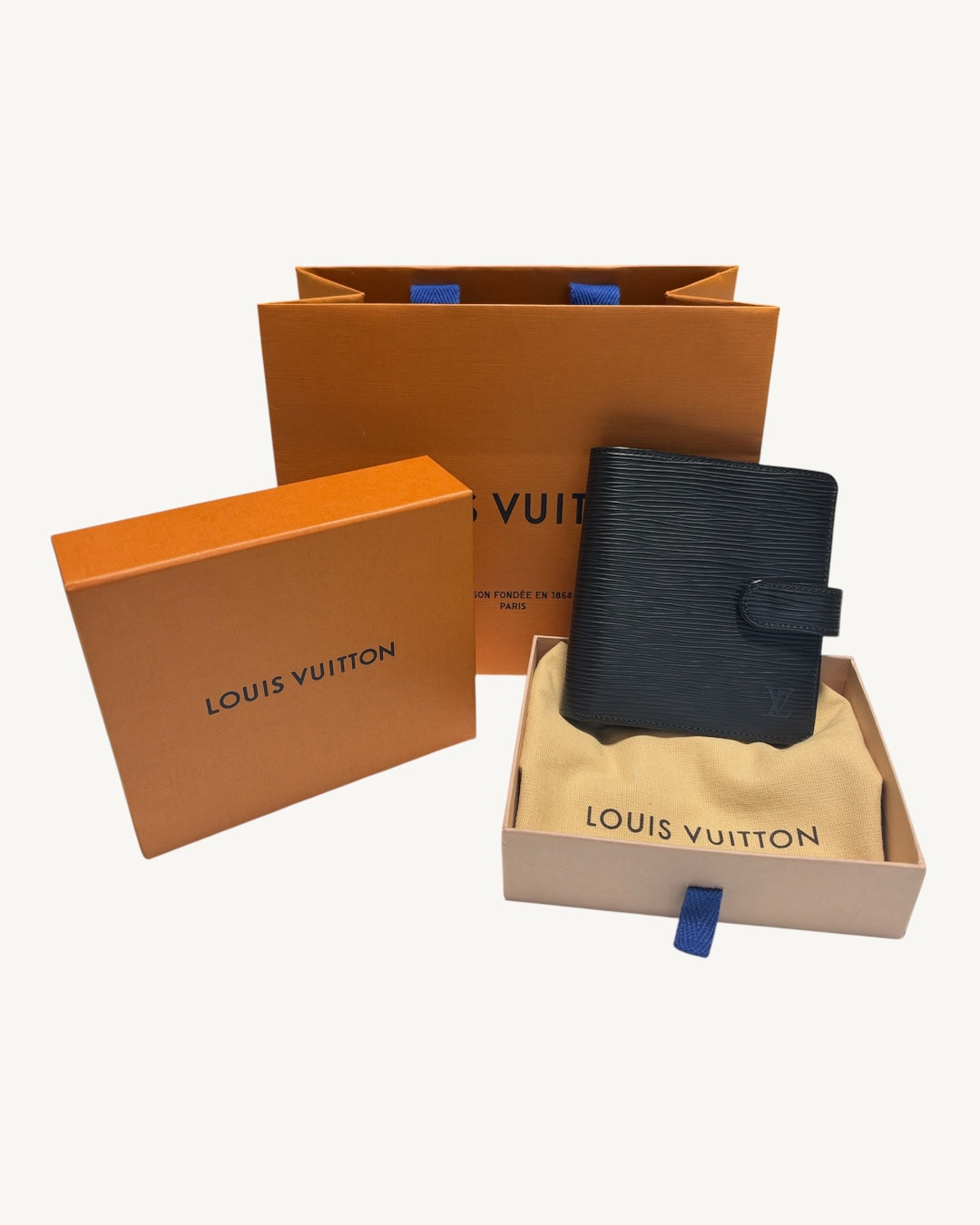 A Louis Vuitton black textured wallet displayed in its original beige dust bag, with the orange Louis Vuitton box lid, shopping bag, and branded dust bag arranged around it on a light gray background.