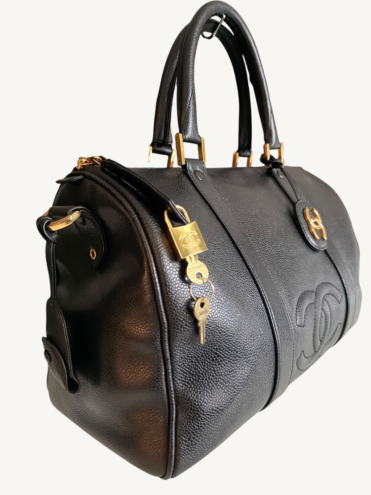 A black pebbled leather duffel bag with gold-tone hardware, featuring a lock and key closure, top handles, and an embossed interlocking CC logo on the side, set against a plain light background.