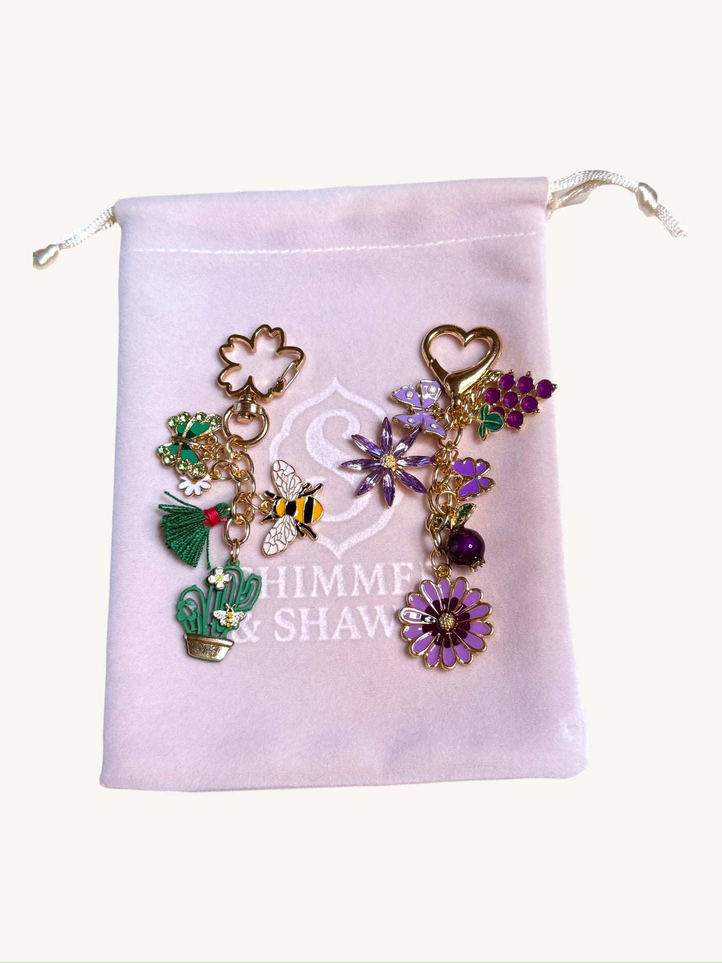 A set of colorful enamel charm keychains displayed on a light pink drawstring pouch. The charms feature gold-tone metal with purple, green, and yellow enamel details, including flowers, a bee, a cactus, and a clover.