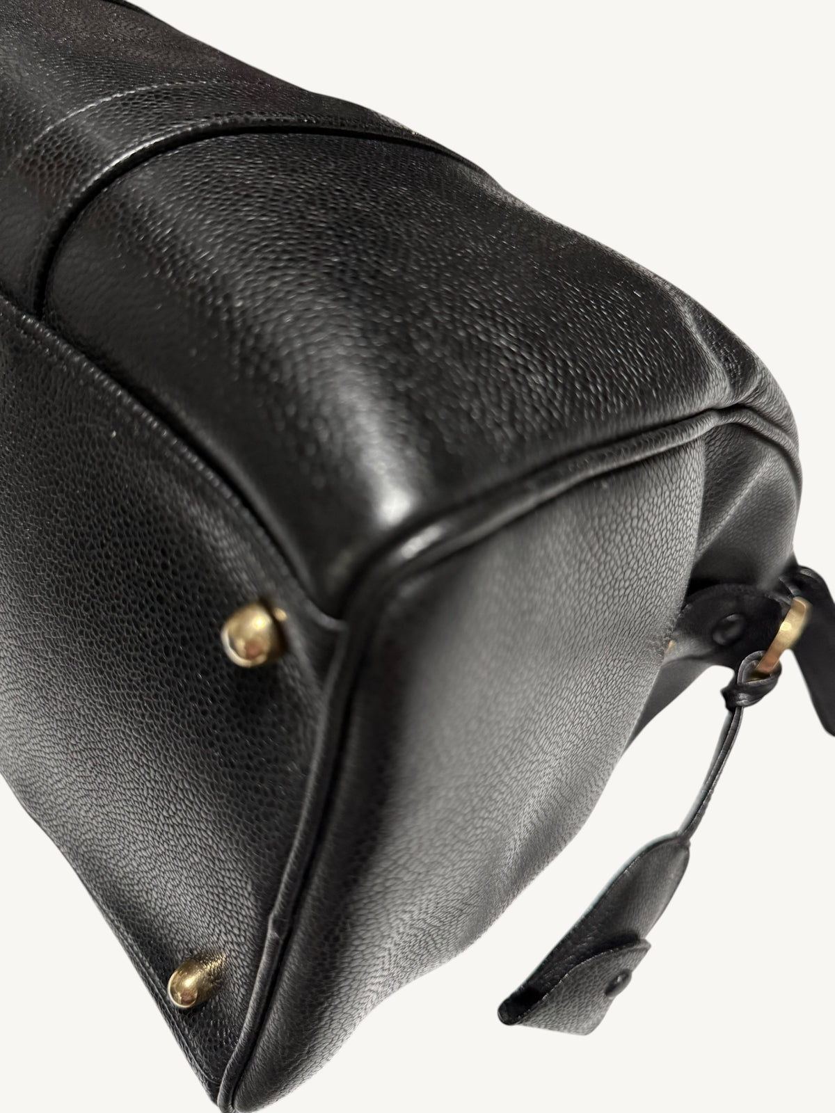 Close-up of a black pebbled leather handbag corner showing a gold-tone metal rivet and a small leather tag on a light off-white background.