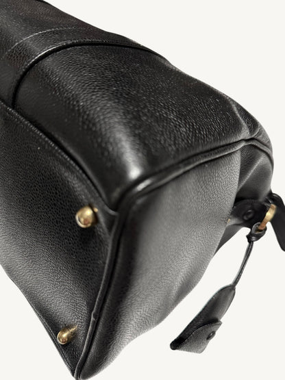 Close-up of a black pebbled leather handbag corner showing a gold-tone metal rivet and a small leather tag on a light off-white background.