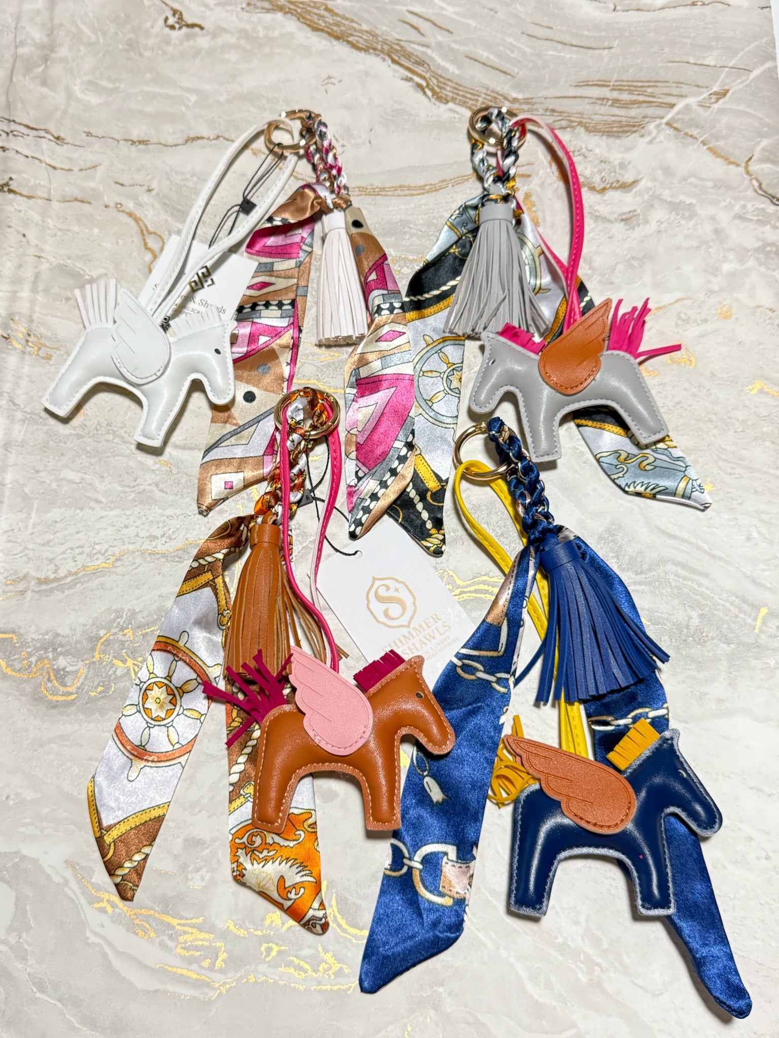 A set of six horse-shaped keychains or bag charms arranged on a light beige marble surface. The set includes various colors and materials: white faux leather, silver metal, brown leather, navy blue velvet, and silk scarves in pink, orange, yellow, and blue.