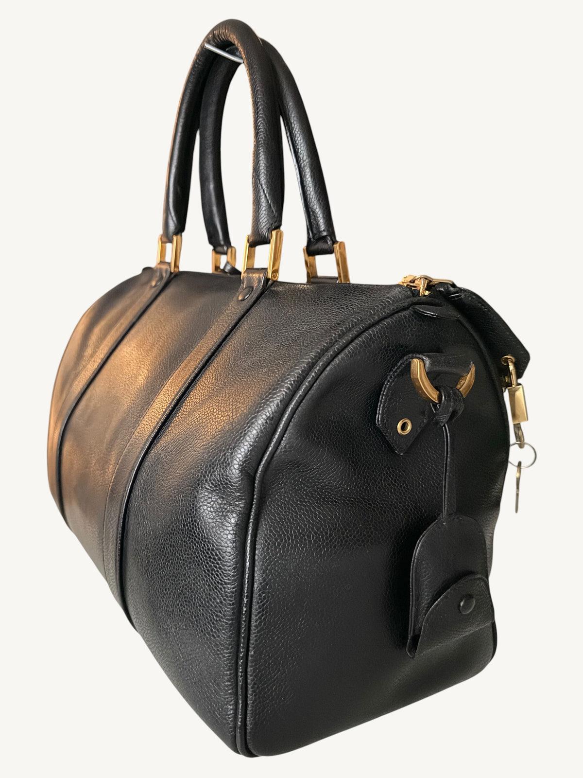 A black pebbled leather CHANEL duffel bag with gold-tone hardware, dual top handles, and a hanging leather tag, shown on a plain light background.