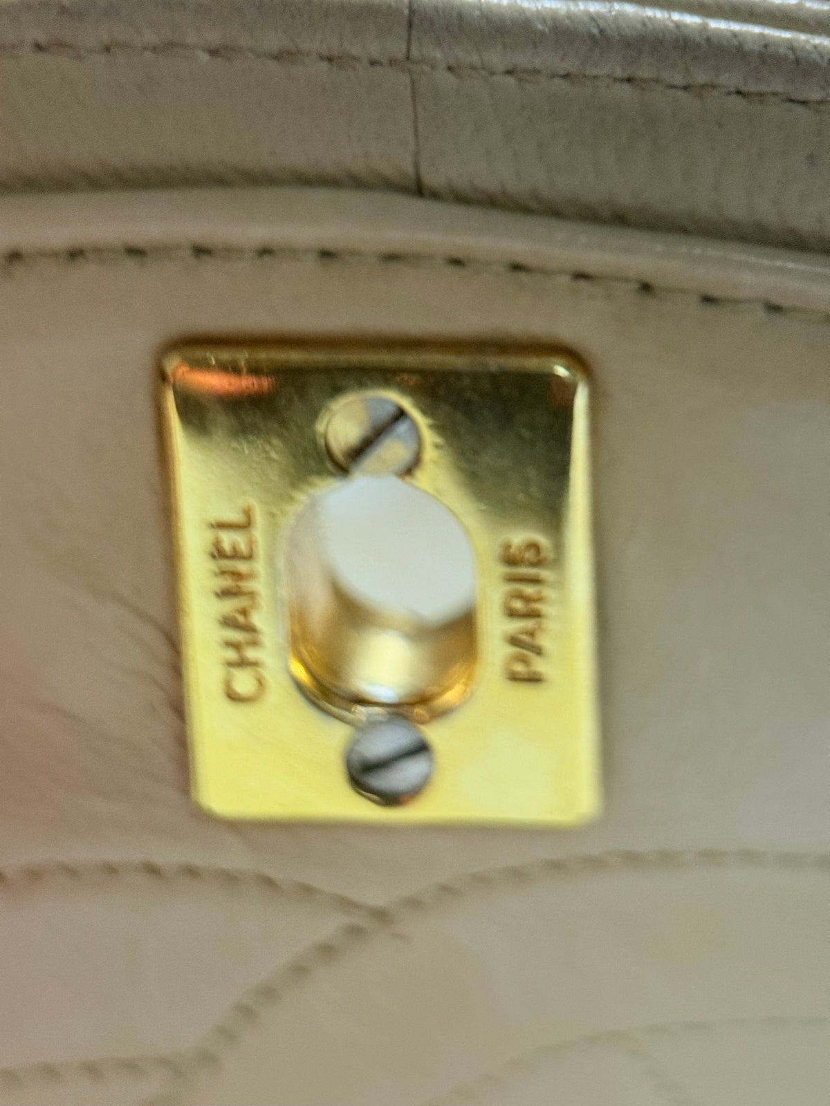 Close-up of a Chanel handbag hardware with a gold-tone metal clasp engraved with 'CHANEL PARIS' on a light beige quilted leather background.