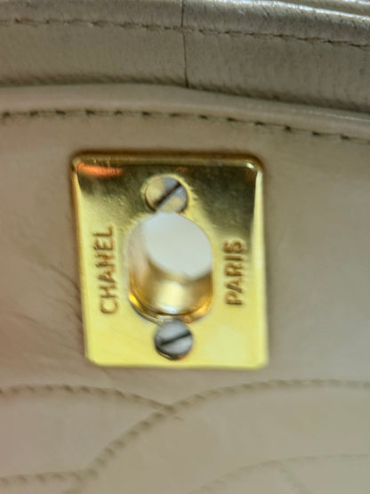 Close-up of a Chanel handbag hardware with a gold-tone metal clasp engraved with 'CHANEL PARIS' on a light beige quilted leather background.