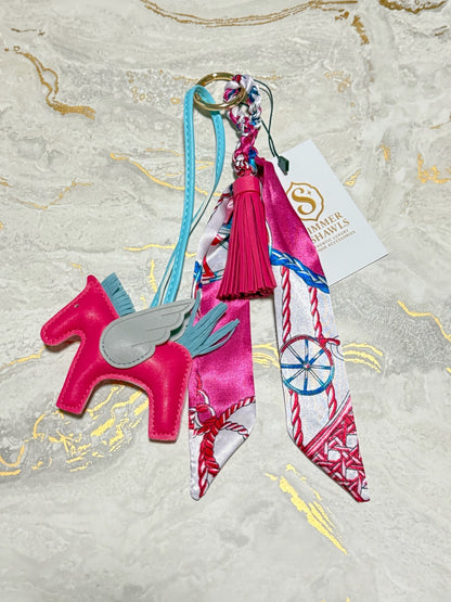 A pink and white decorative keychain or bag charm set featuring a leather horse with a wing, patterned silk ribbon tails, a pink tassel, and a branded paper tag on a light marble-like surface.