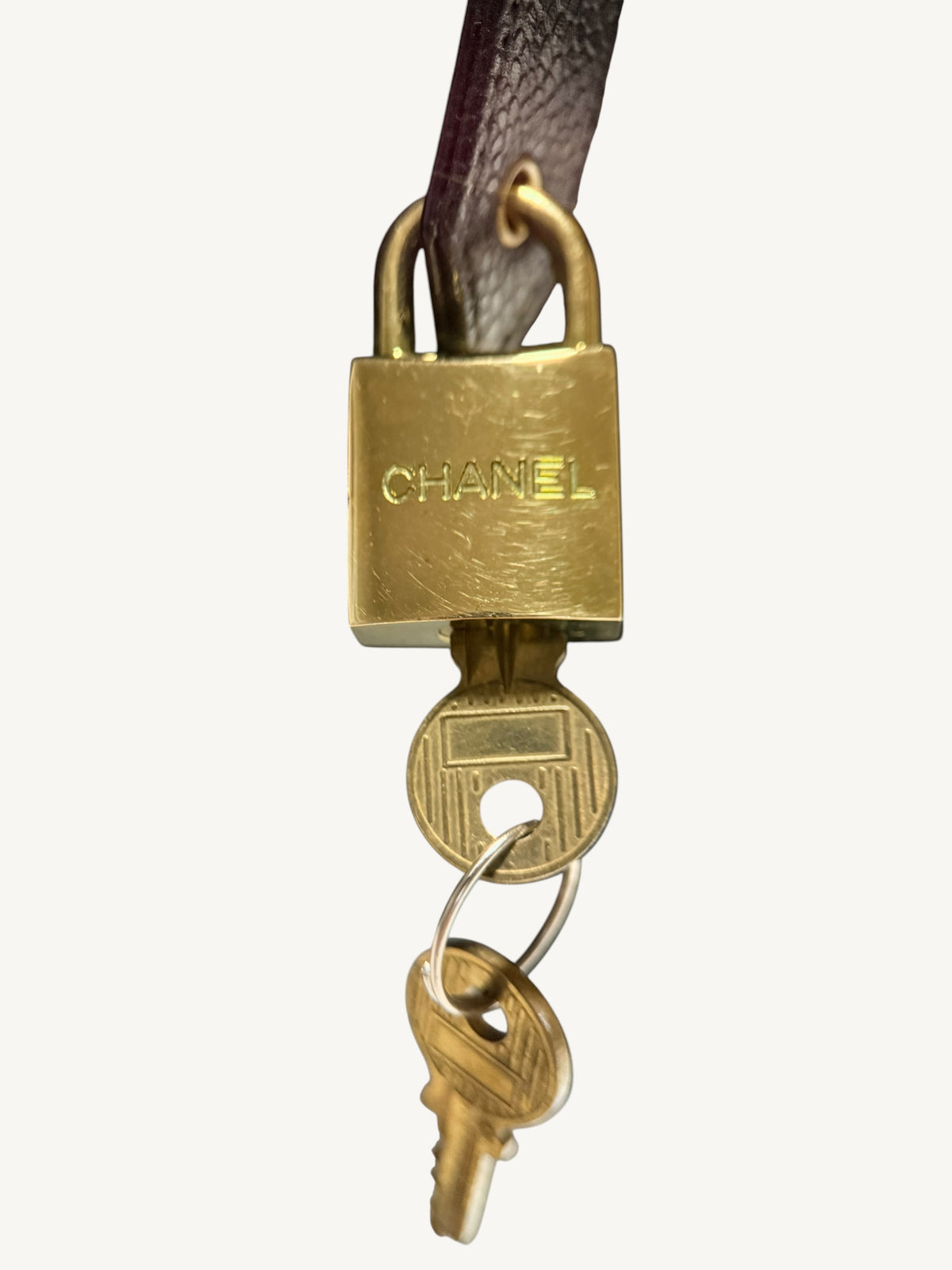 A Chanel padlock with a leather strap and two keys, shown against a plain light background. The padlock is engraved with the CHANEL logo.