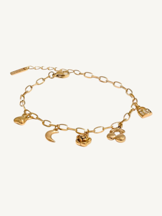 A gold chain bracelet with multiple dangling charms, including a crescent moon, rose, cloud, and small heart, displayed on a light cream background.