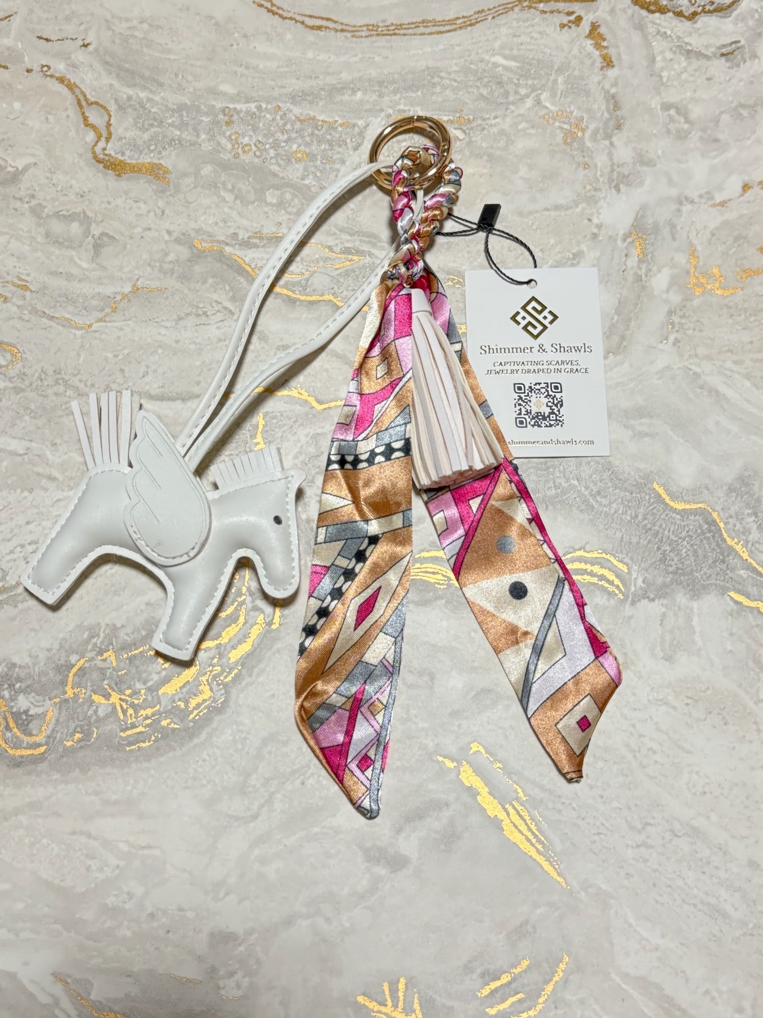 A decorative keychain or bag charm laid flat on a light marble-like surface with gold veining. The charm includes a white horse-shaped tag, a gold key ring, a braided strap, a cream tassel, and a patterned fabric ribbon in pink, beige, brown, black, and white.