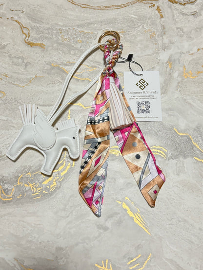 A decorative keychain or bag charm laid flat on a light marble-like surface with gold veining. The charm includes a white horse-shaped tag, a gold key ring, a braided strap, a cream tassel, and a patterned fabric ribbon in pink, beige, brown, black, and white.