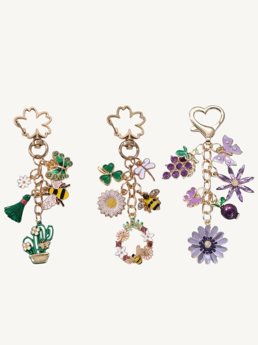 Three gold-tone charm earrings displayed side by side on a light cream background. Each earring features a floral-shaped top clasp and dangling charms: the left has a shamrock, green leaves, a bumblebee, and a cactus; the middle has a shamrock, a daisy, a bumblebee, and a small wreath; the right has purple grapes, a purple flower, a purple heart, and a purple daisy.