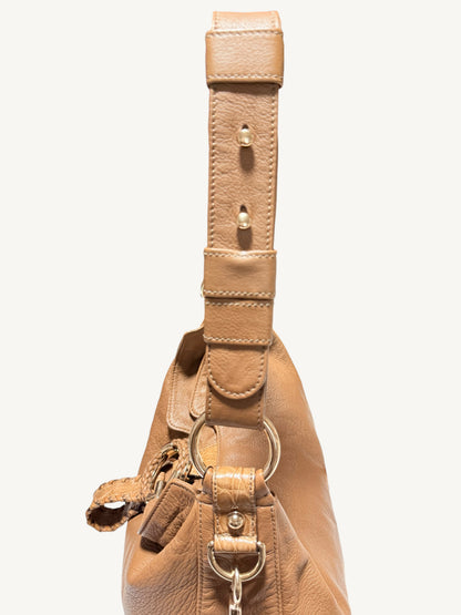 Close-up of a brown leather shoulder bag showing the strap, metal hardware, and a small hanging tag against a plain light background.