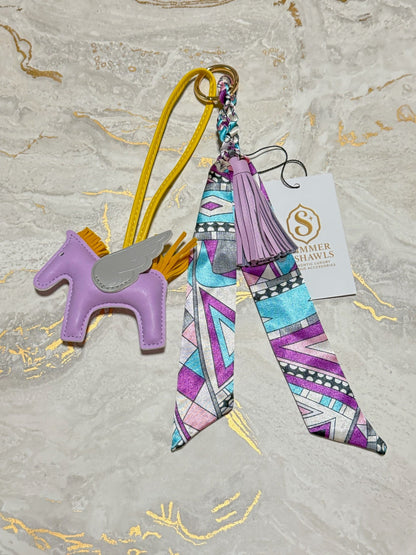A handmade keychain or bag charm featuring a purple faux-leather unicorn with a silver wing and yellow strap, alongside a colorful geometric patterned ribbon with a purple tassel and a Summer Travels brand tag, displayed on a light marble background.