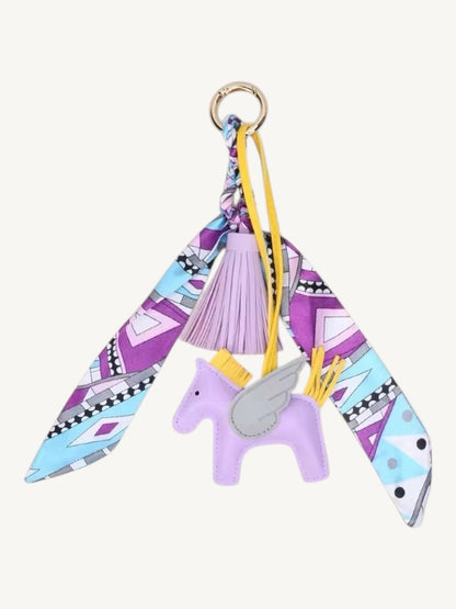 A whimsical keychain charm featuring a light purple leather unicorn with a gray wing, a lavender tassel, and a split scarf with a purple, blue, and white geometric pattern, hanging from a gold ring.