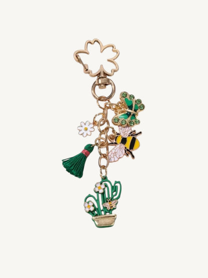 A gold-tone metal keychain charm featuring a clover-shaped ring, a green and yellow bumblebee, a green leafy sprig, a small white flower, a green tassel, and a potted cactus with white daisies, all on a light cream background.