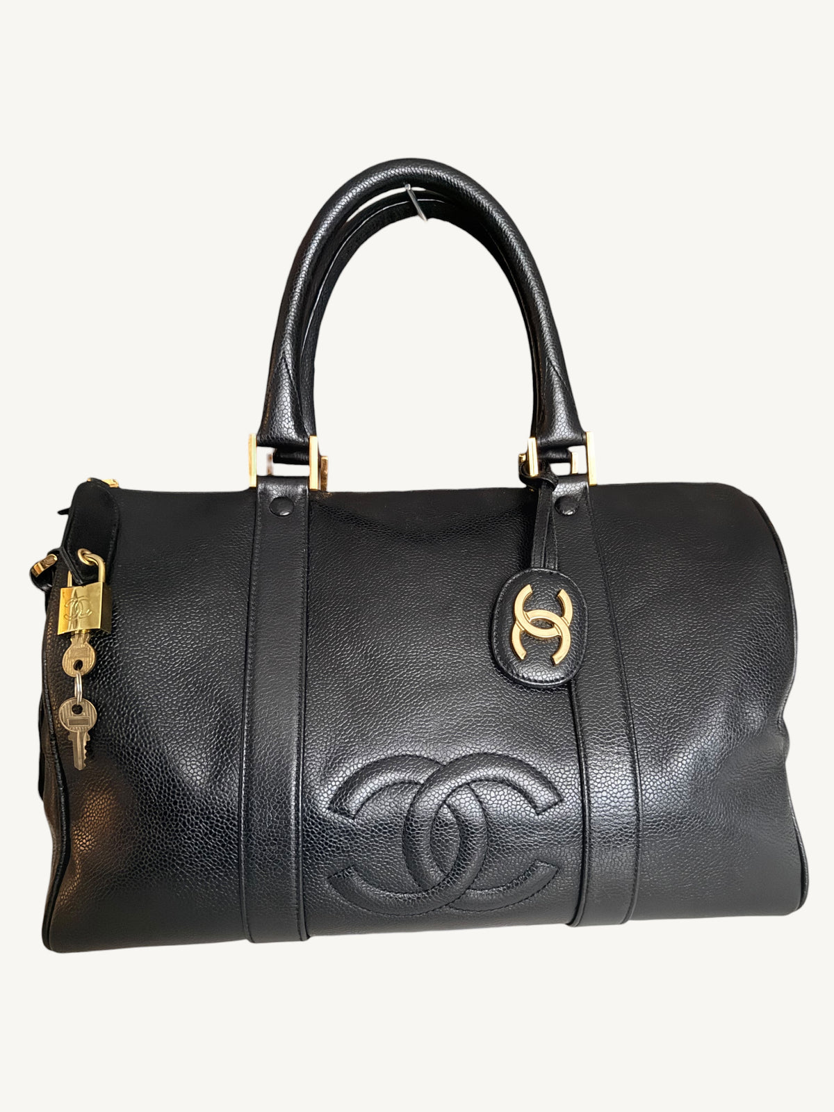 A black leather Chanel duffle bag with gold-tone hardware, featuring the embossed Chanel logo on the front, a top handle, and a lock with keys hanging from the side.